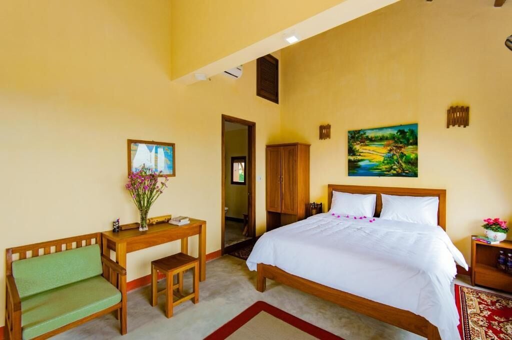 A bedroom with a bed, a wooden desk, a green bench, a painting, and a wardrobe.