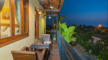 A balcony with wooden furniture and a table, overlooking a garden with various plants and a view of the city at dusk.