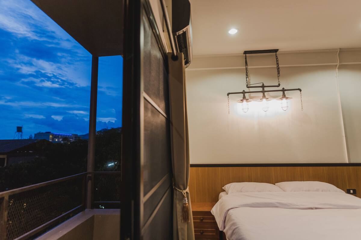 A hotel room with a bed, a nightstand, a hanging light fixture, and a view of the sky.