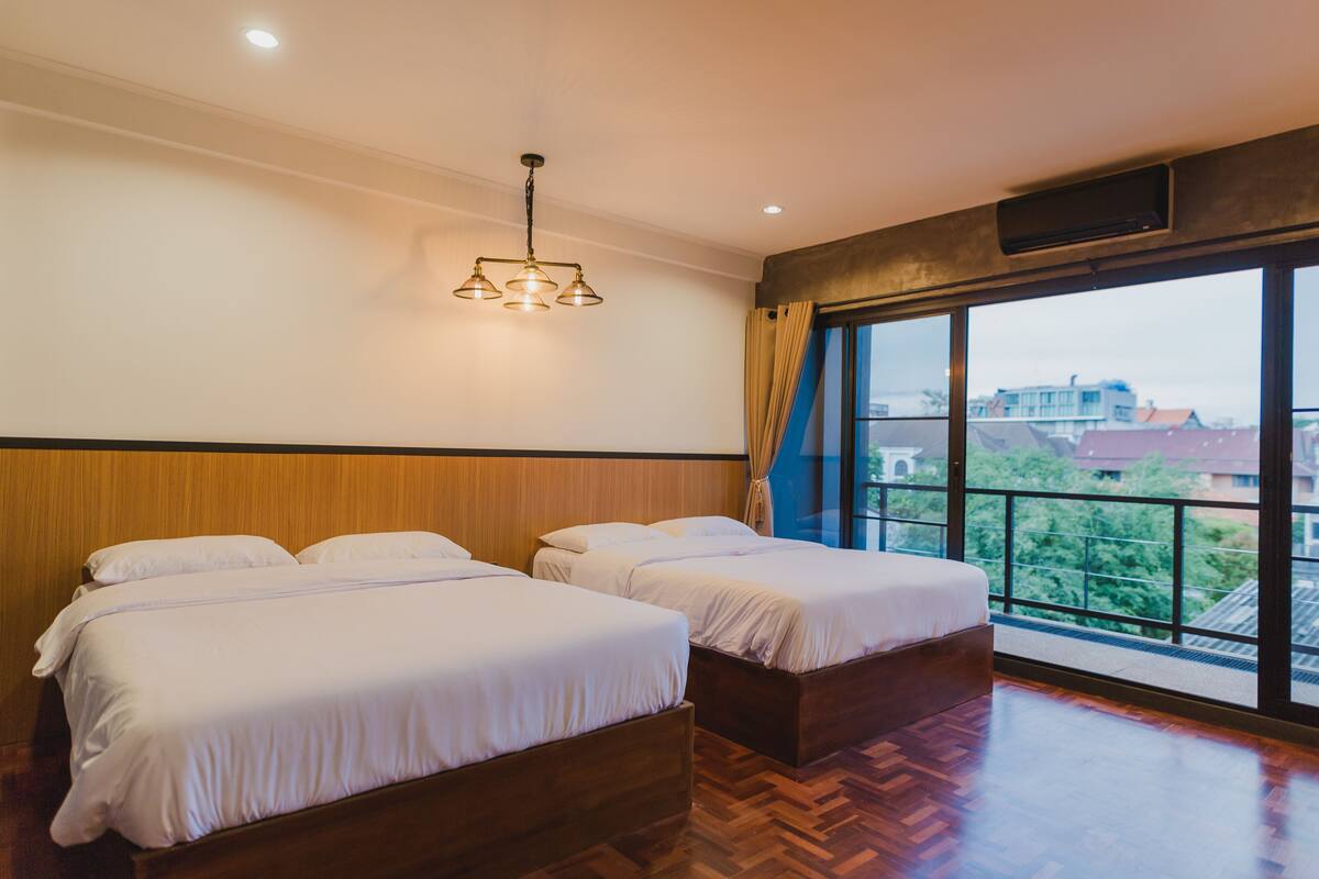 A hotel room with two beds, a wooden floor, a large window with a view, and a ceiling light fixture.