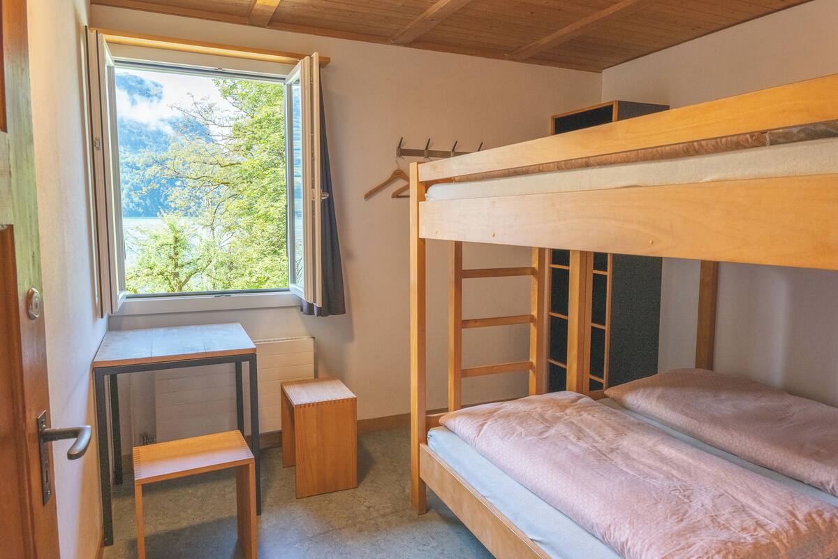 A bunk bed with a desk and a small stool in a room with a window overlooking greenery.