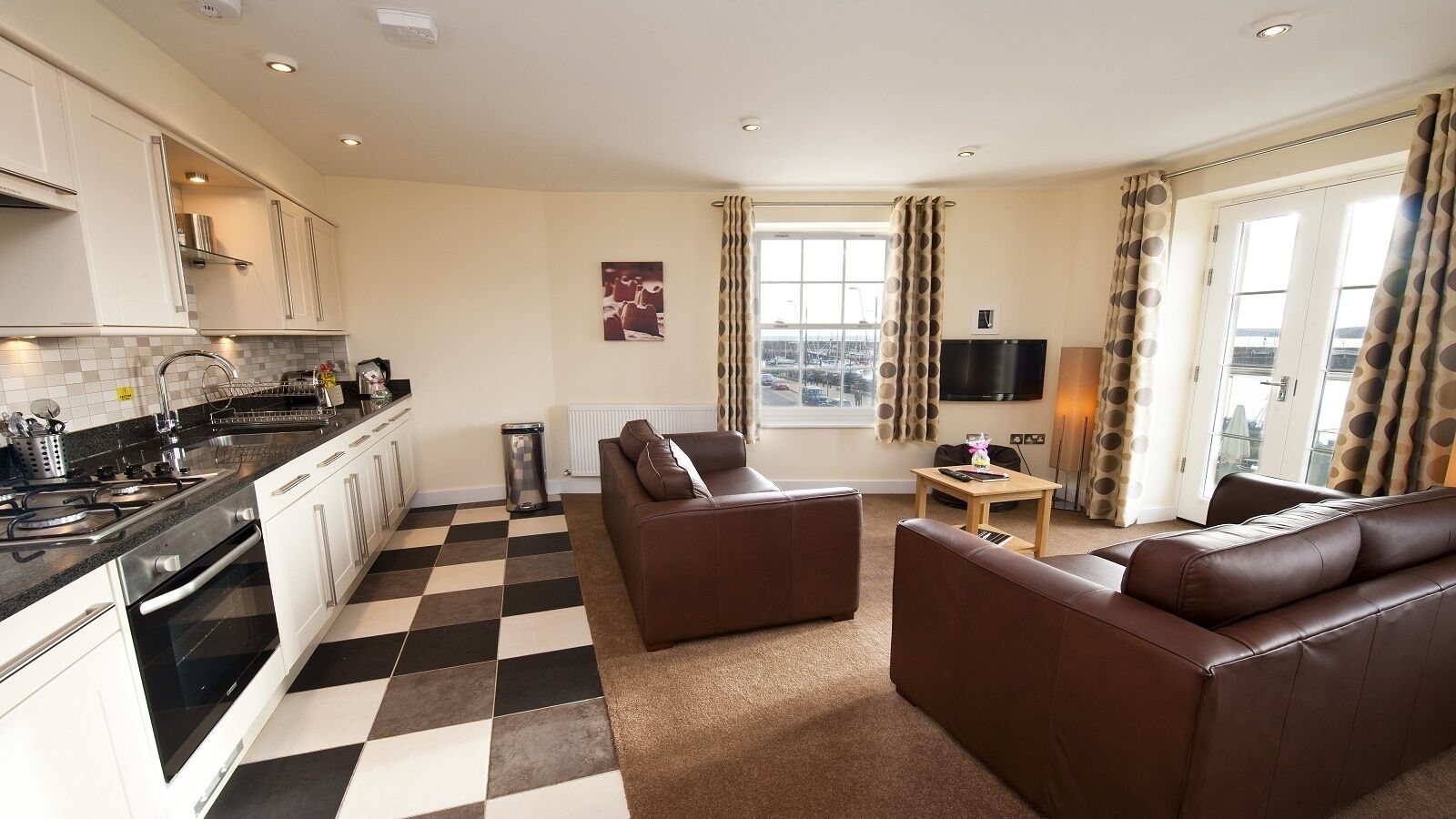 A modern kitchen with a black and white checkered floor, a brown leather sofa, and a flat-screen TV.