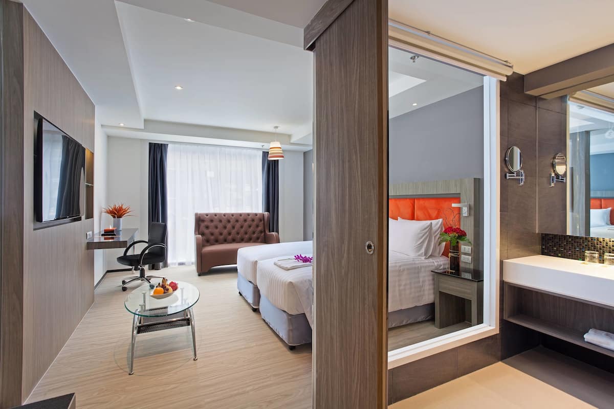 Une chambre d’hôtel moderne, équipée d’un lit, d’une table de chevet et d’un grand miroir. On y trouve également un coin salon avec un fauteuil et une petite table.
