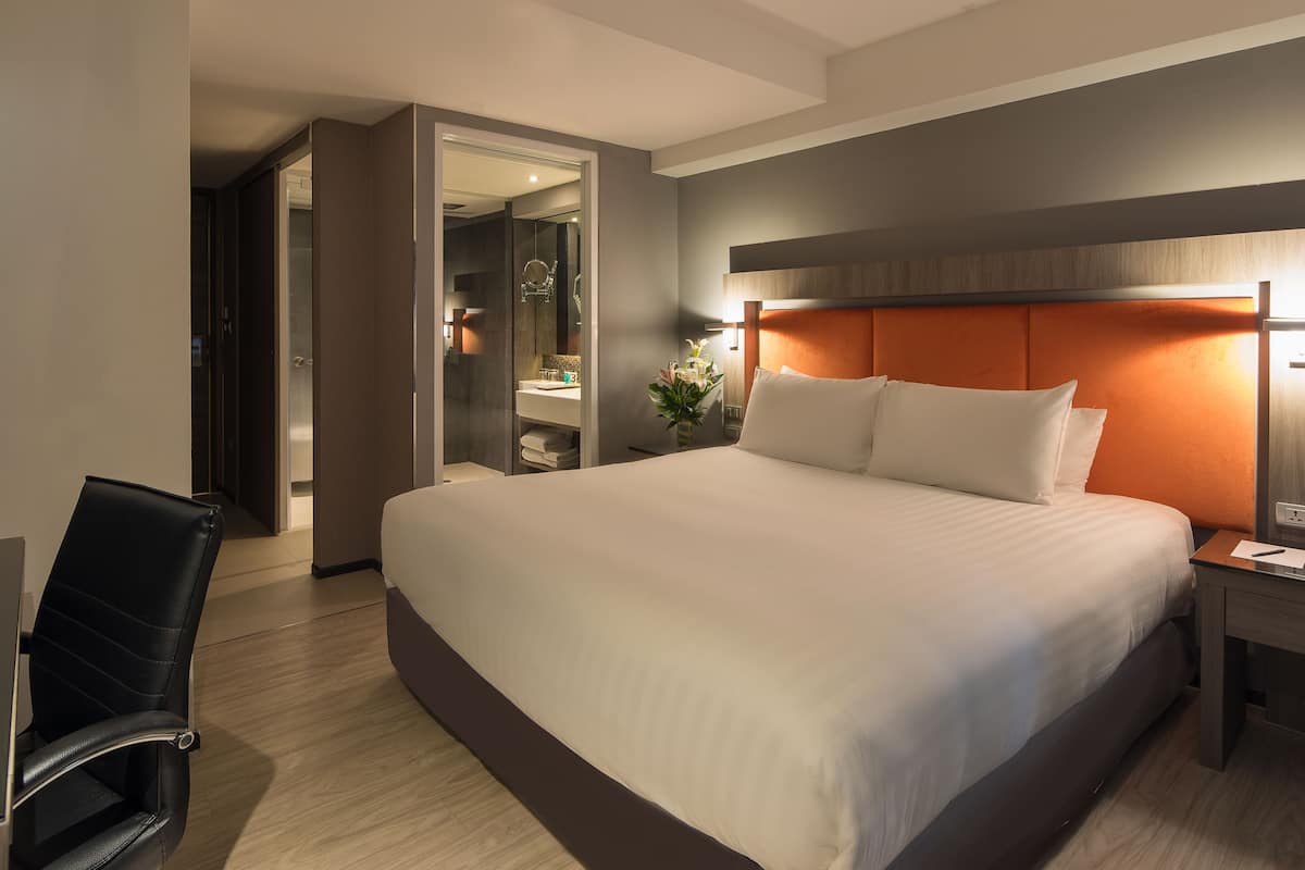 Une chambre d’hôtel avec un grand lit, un bureau, une chaise et une salle de bain visible à travers une porte ouverte.