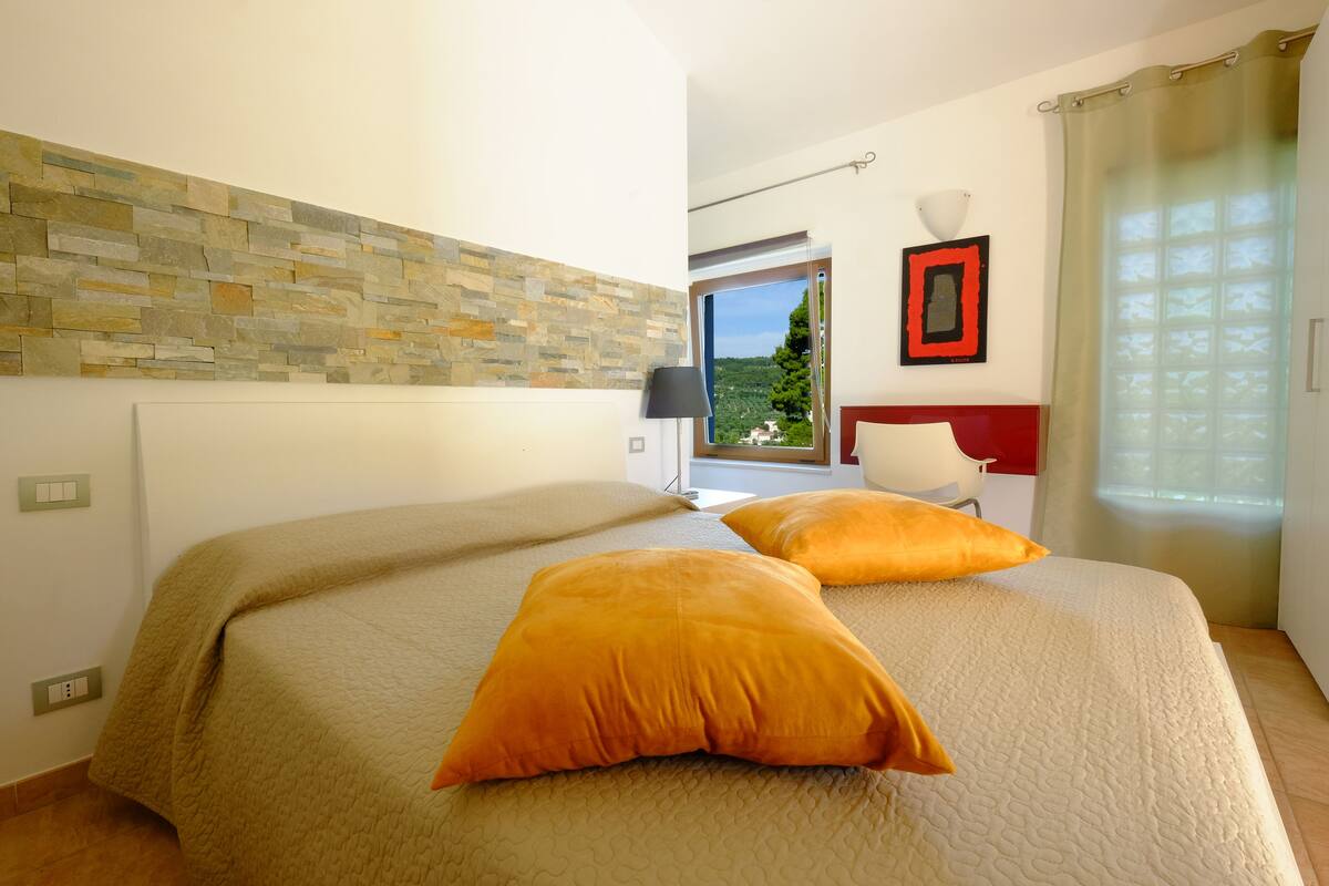 A bedroom with a stone wall, a bed with two orange pillows, a nightstand, a lamp, and a window with a view of greenery.