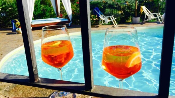 Deux verres d’une boisson rouge, posés au bord d’une piscine, avec vue sur la piscine, des chaises longues et une pergola.