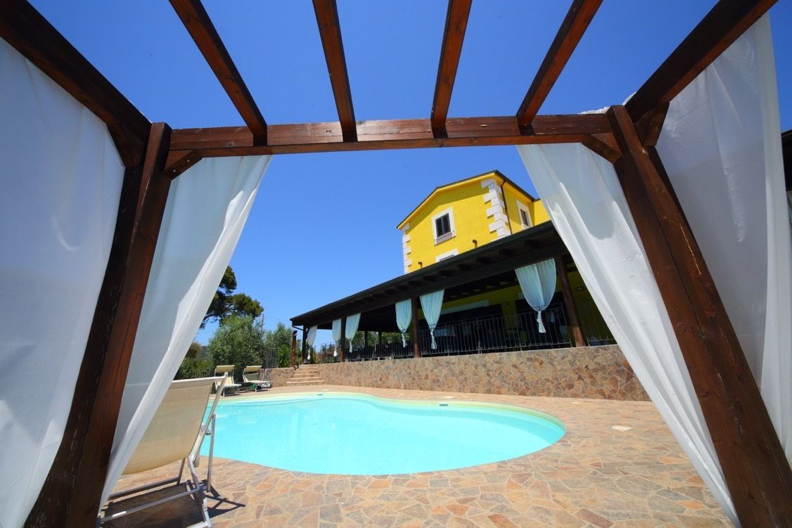 A swimming pool with a stone patio, a yellow building in the background, and a wooden pergola structure.