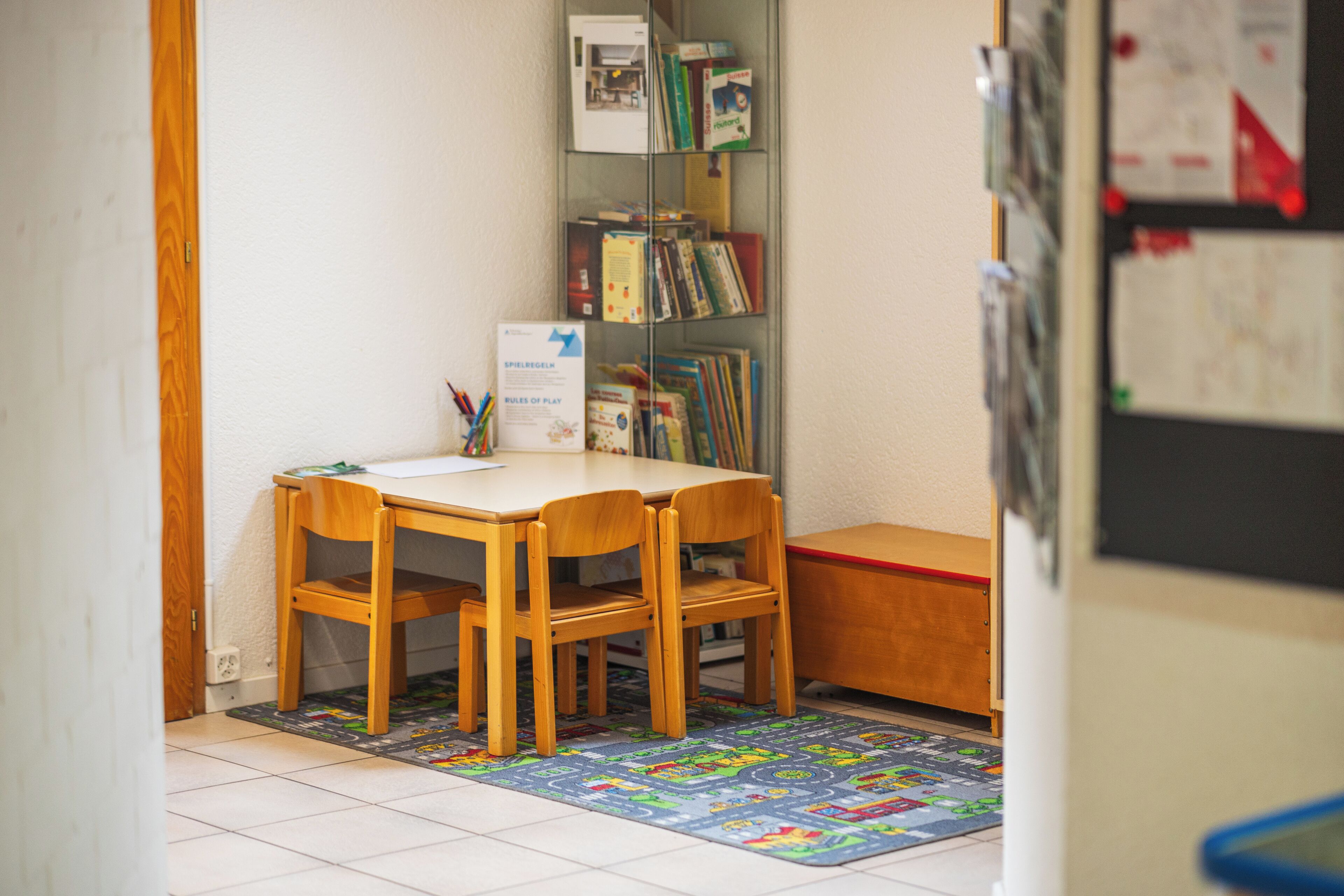 Une petite pièce, bien rangée, avec une table et des chaises en bois, une bibliothèque remplie de livres et un tapis coloré sur le sol.