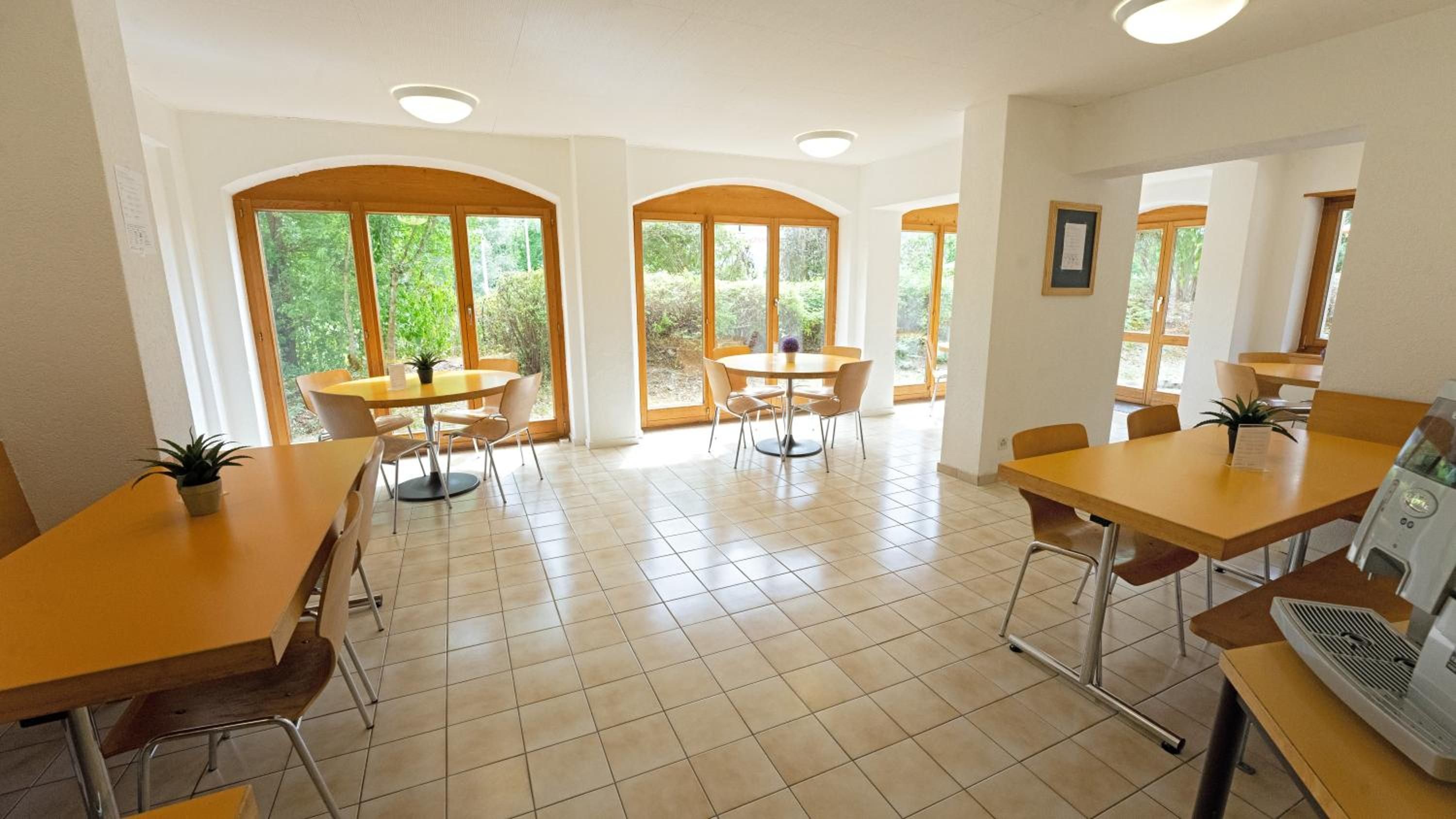 A dining area with multiple tables and chairs, large windows with natural light, and a tiled floor.