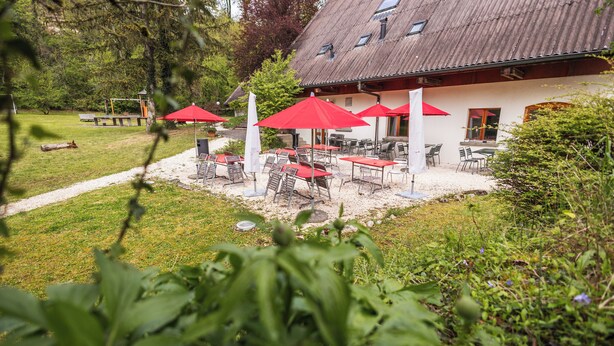 Ein Sitzbereich im Freien mit roten Sonnenschirmen, ein weißes Gebäude mit geneigtem Dach und eine Rasenfläche.