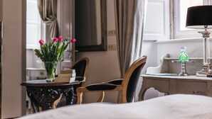 Une chambre d’hôtel avec un lit, une petite table ronde ornée d’un vase de fleurs, deux chaises, un bureau avec une lampe et une fenêtre avec des rideaux.