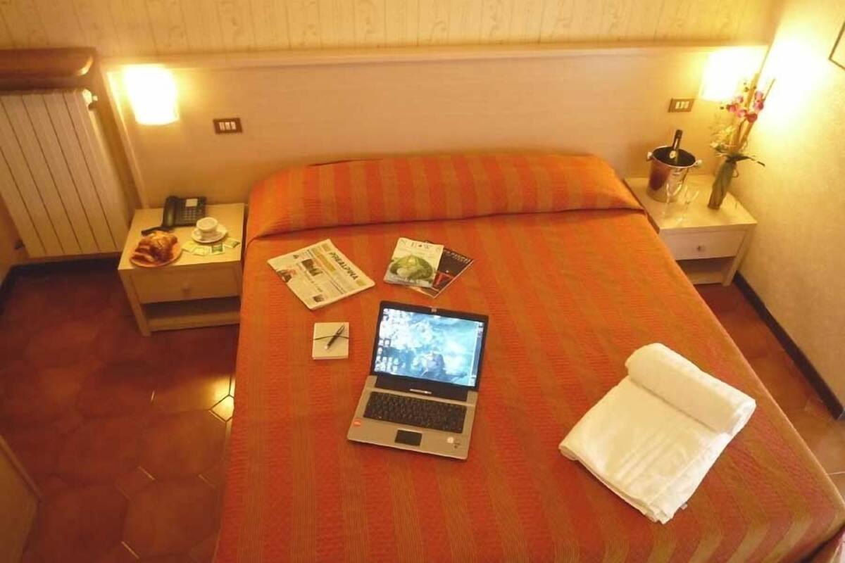 A hotel room with a bed, a laptop, a nightstand, and a bedside table with a lamp and a vase of flowers.