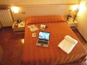 A hotel room with a bed, a laptop, a nightstand, and a bedside table with a lamp and a vase of flowers. - Hotel Montecarlo (Castellanza)