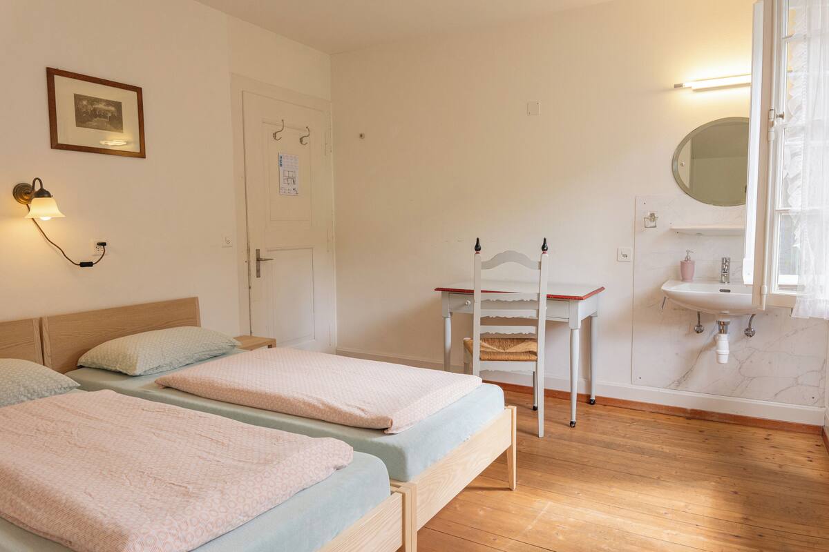 Ein Schlafzimmer mit zwei Betten, einem kleinen Tisch, einem Spiegel und einem Badezimmer mit Waschbecken und Spiegel.