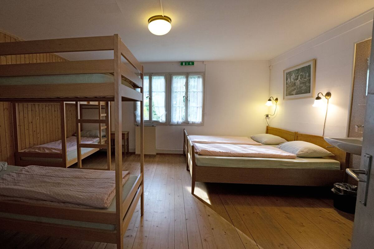 Ein Schlafsaalzimmer mit Etagenbetten, einem Einzelbett, einem Fenster mit Vorhängen, einer Wandleuchte und einem gerahmten Bild.