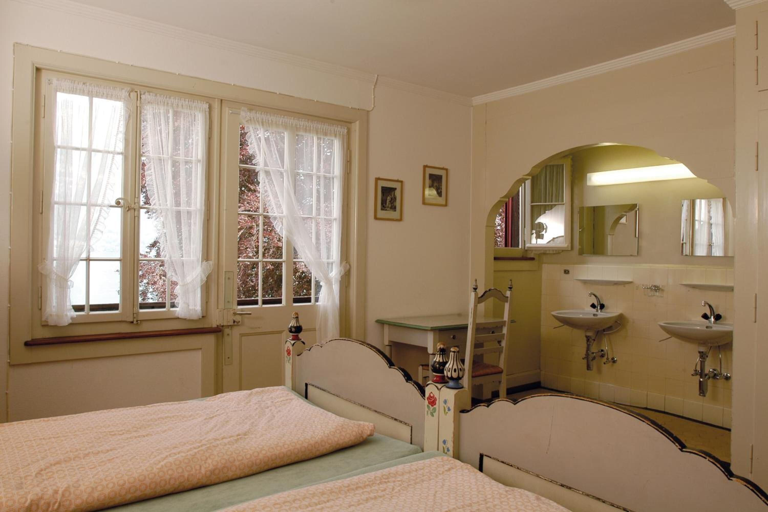 Ein Schlafzimmer mit zwei Betten, einem Fenster mit Vorhängen, einem Schminktisch mit Spiegel und einem Waschbecken.