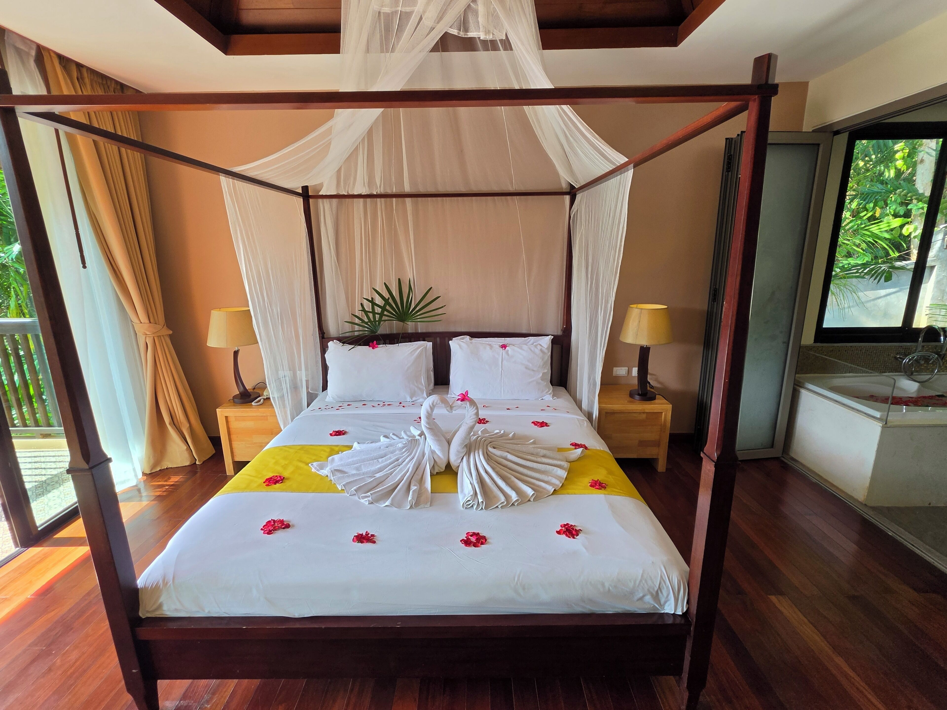 A four-poster bed with a canopy, two bedside lamps, a bedside table, and a view of the outdoors.