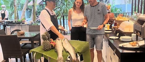 A restaurant with a tropical theme, featuring a chef preparing fish on a mobile serving cart, a dining area with wicker chairs, and a counter with food items.