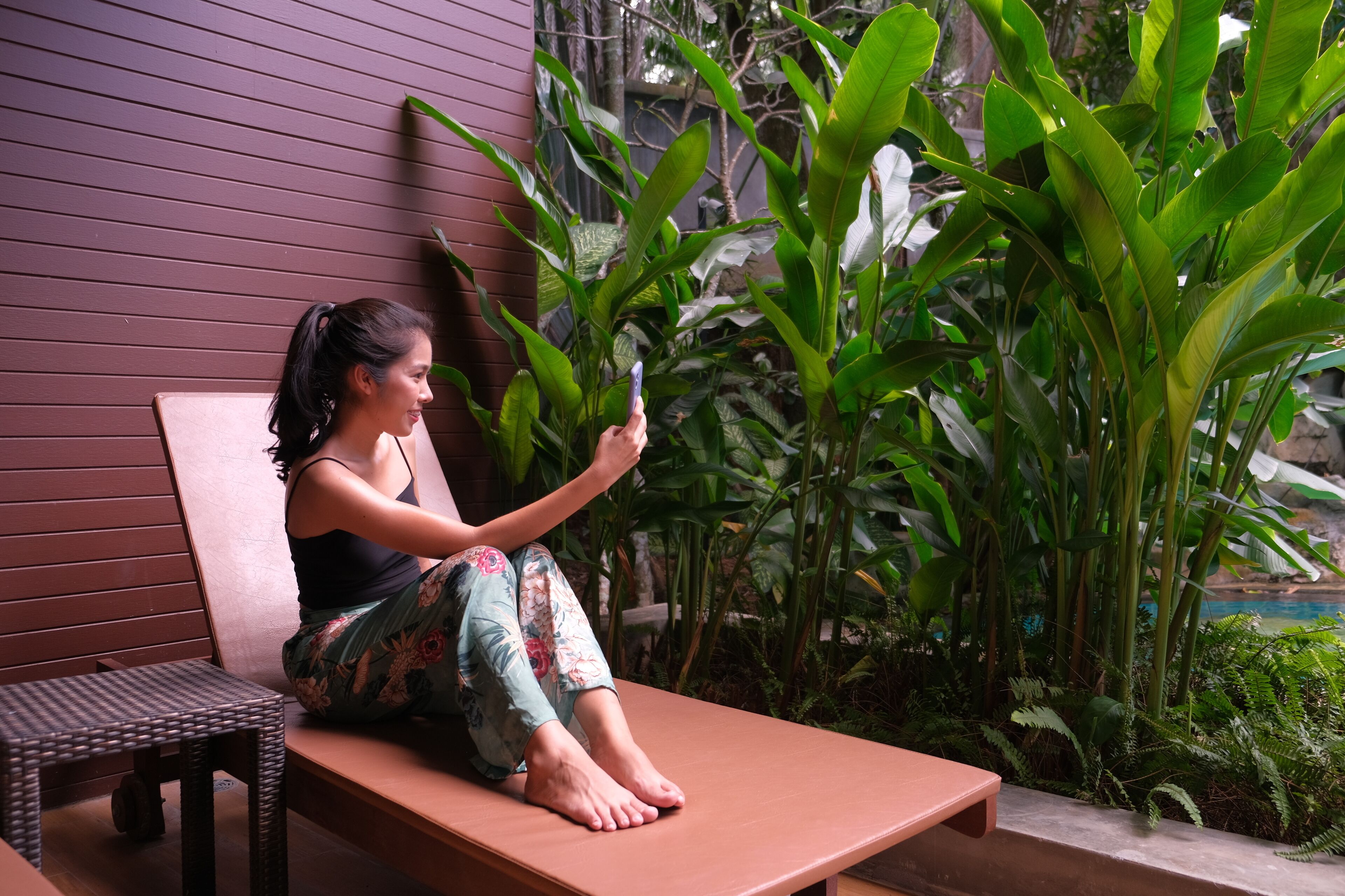Une personne assise sur un banc en bois, à l’extérieur, avec une végétation luxuriante et une piscine en arrière-plan.