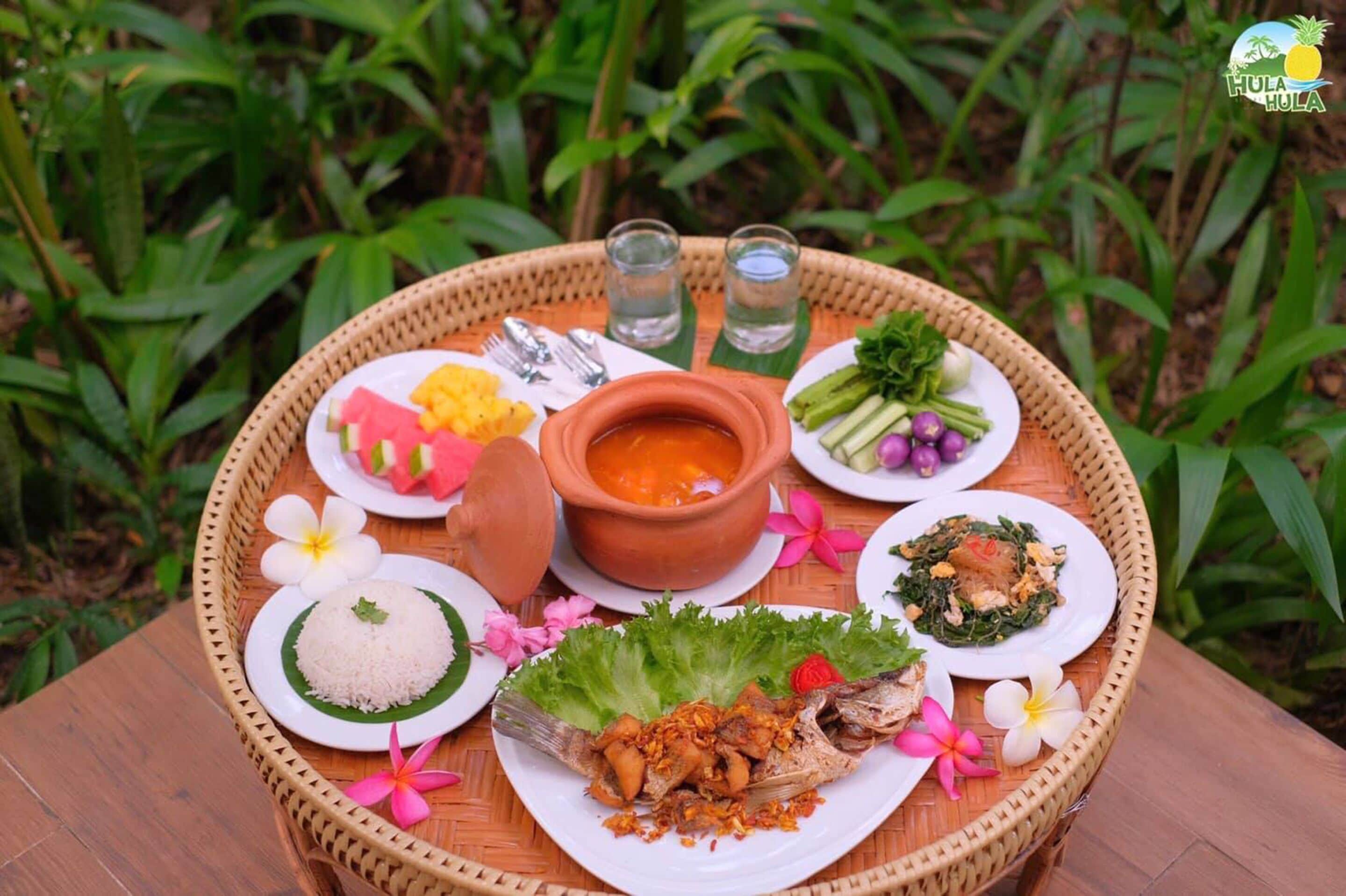 A round wooden tray with various dishes, including rice, soup, and grilled fish, set on a table outdoors.