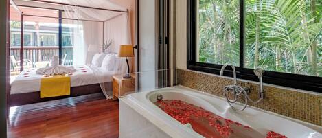 A bedroom with a large bed, a bathtub with rose petals, a window with a view of palm trees, and a balcony with chairs.