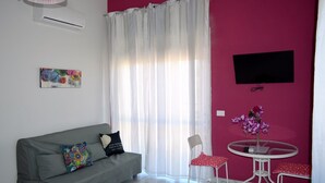 Un salon avec un canapé gris, un fauteuil blanc, une petite table ronde ornée d’un vase de fleurs et un téléviseur fixé sur un mur rose.