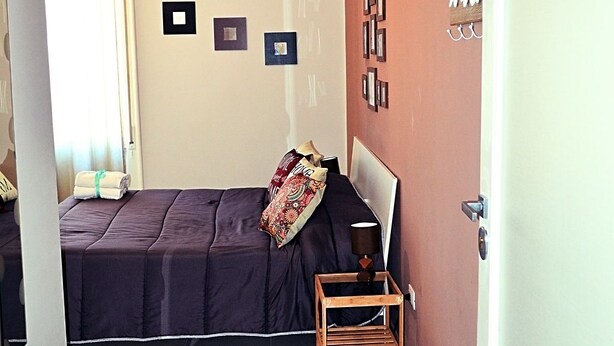 A small, neatly arranged bedroom with a bed, a bedside table with a lamp, a wall-mounted air conditioner, and a door with a number sign. - Nel Cuore di Parthenope (Naples)