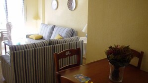 A cozy living area with a striped sofa, a wooden dining table with a vase of flowers, and two wall decorations. - Isla Grosa Apartamentos (San Javier)