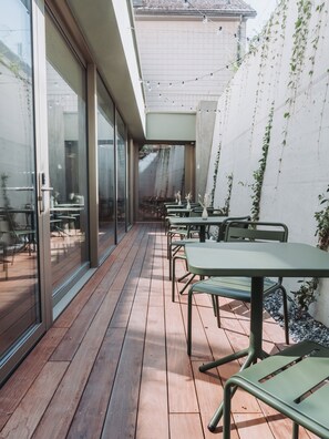 Un patio al aire libre con suelo de madera, mesas y sillas de metal verde y vista a un edificio con puerta de vidrio.