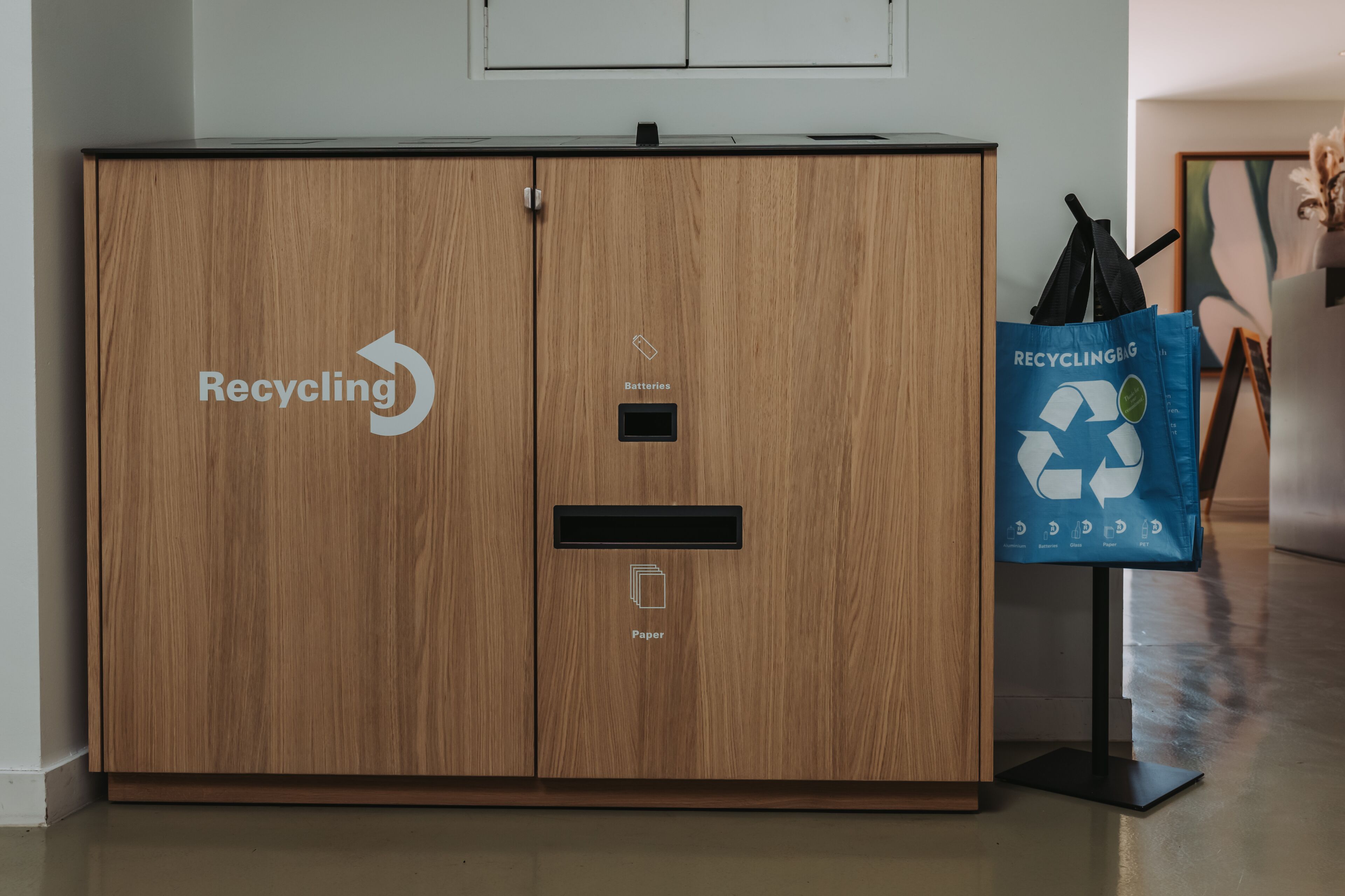 A wooden cabinet with recycling bins for batteries and paper, and a recycling bag on a stand.