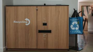 A wooden cabinet with recycling bins for batteries and paper, and a recycling bag on a stand.