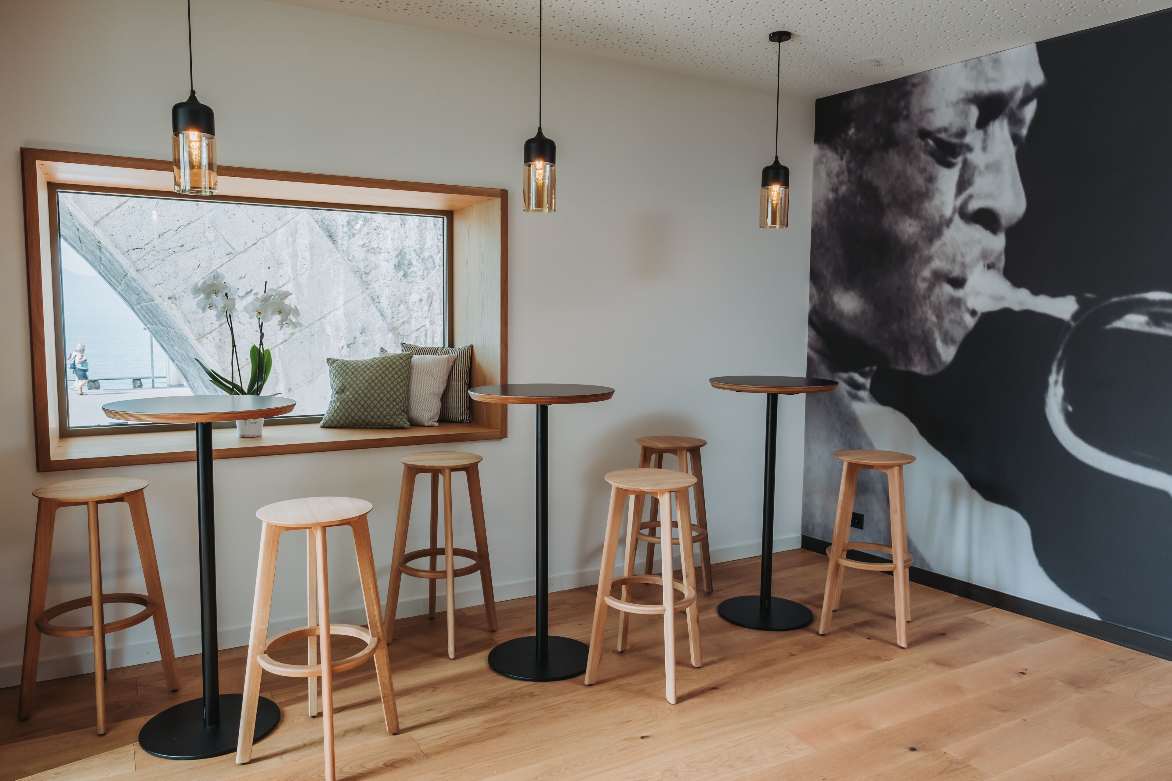 Un interior moderno con piso de madera, mesas redondas y taburetes. Se observa un ventanal con vistas a un paisaje urbano.