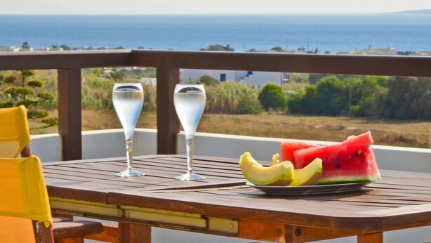A wooden table with a plate of watermelon and bananas, two glasses, and a view of the sea. - Anthi Studios (Naxos)