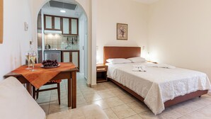 A hotel room with a bed, a small table with a fruit bowl and glasses, a nightstand, and a view of the kitchen through an archway. - Anthi Studios (Naxos)