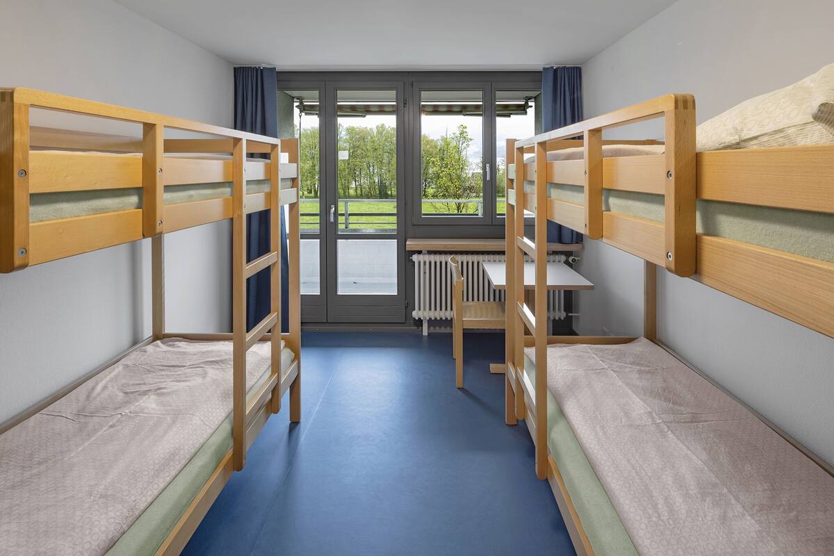 A dormitory room with bunk beds, a desk, and a view of the outdoors.