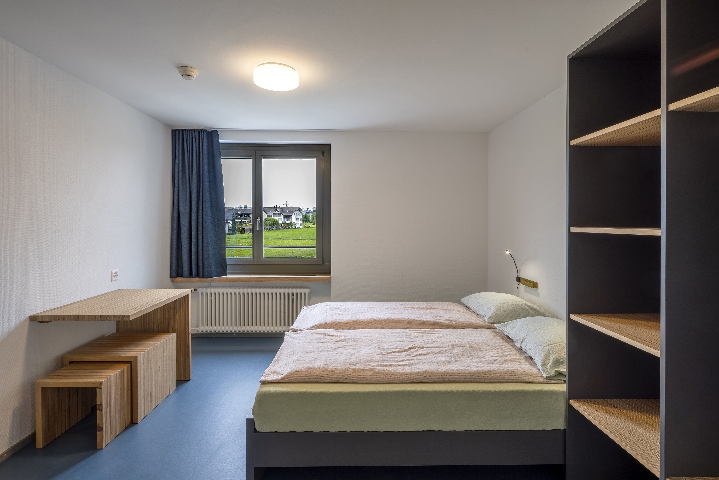 Un dormitorio con cama, escritorio y ventana con vistas a casas.