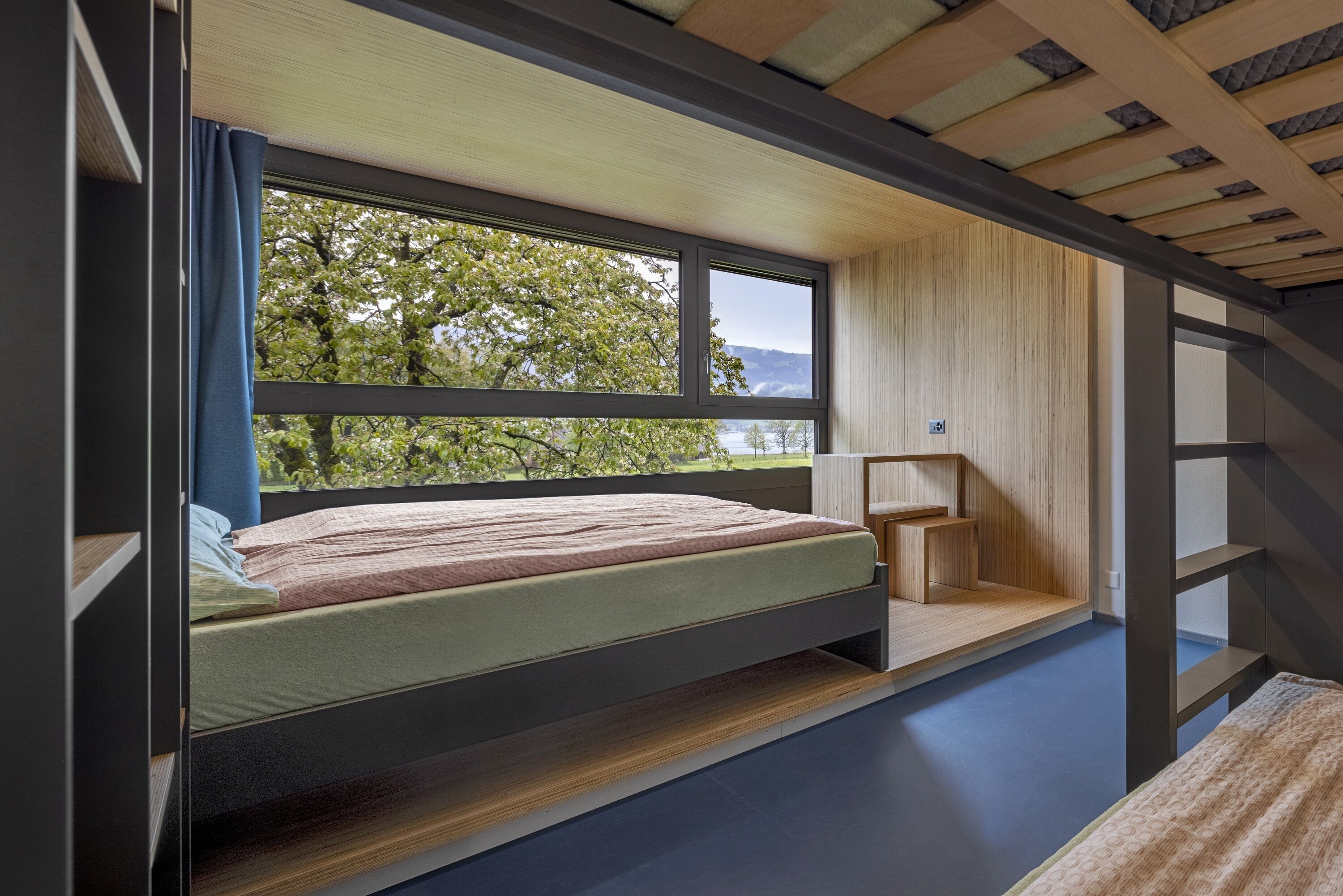 Un dormitorio moderno con cama empotrada, mesita de noche pequeña y vistas a árboles y montañas a través de amplios ventanales.