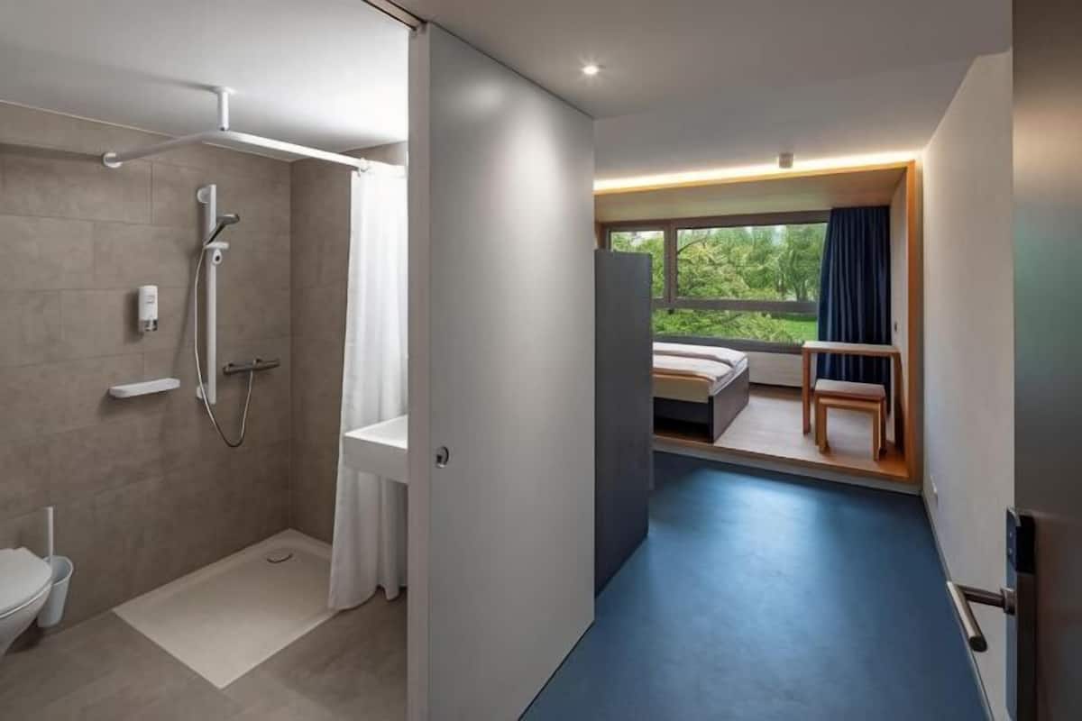 A modern bathroom with a shower, toilet, and a view of a bedroom with a bed and a chair.