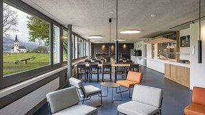 A modern cafeteria with tables and chairs, large windows with a view of a church and mountains, and a counter with a menu board. - Youth Hostel Rapperswil-Jona (Rapperswil-Jona)