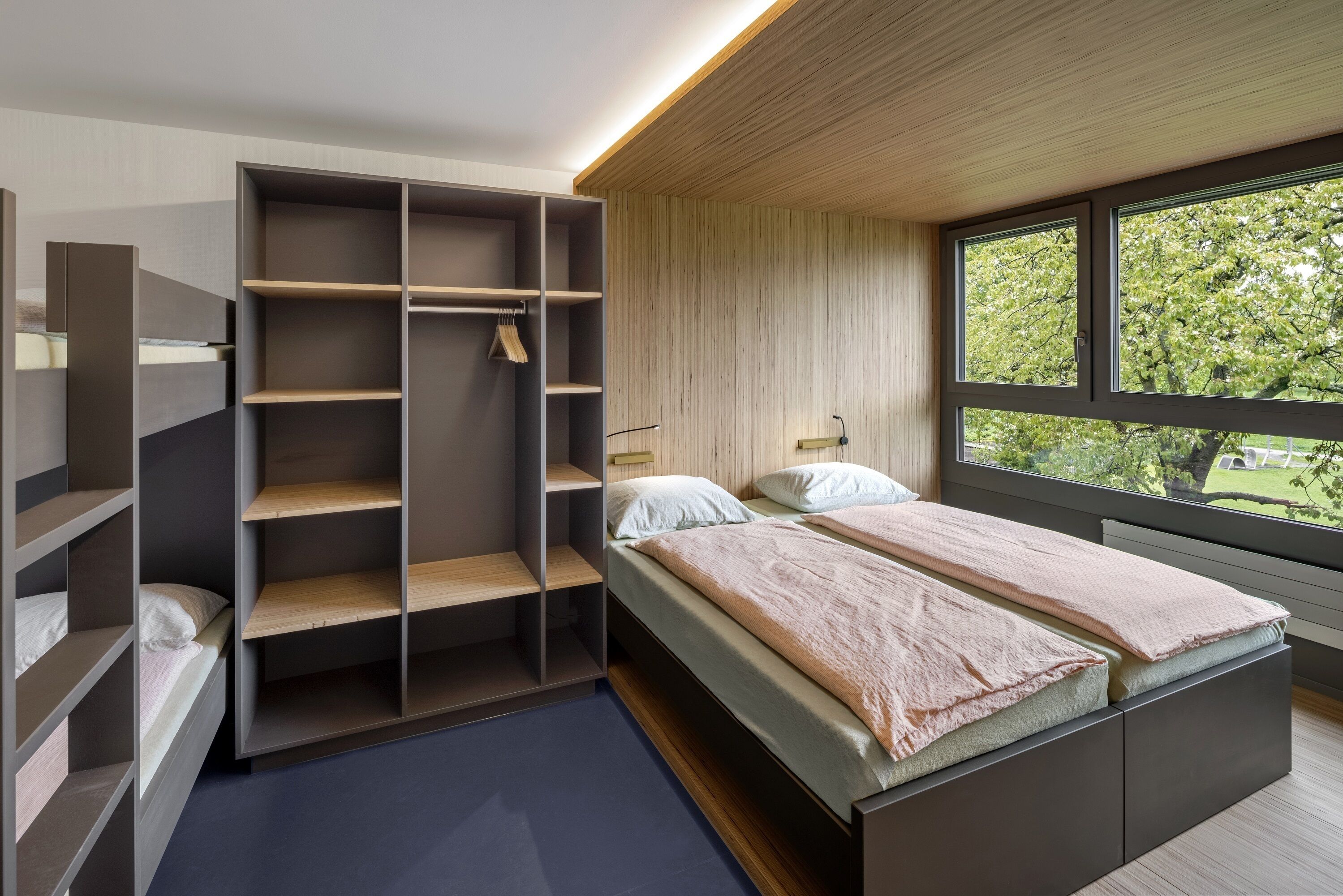 A modern bedroom with a built-in wardrobe, a large window with a view of trees, and a bed with a wooden headboard.