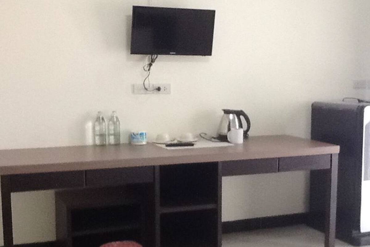 Una habitación con una televisión de pantalla plana montada en la pared, un escritorio con hervidor, tazas y botellas, y una maleta en el suelo.