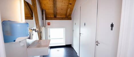 A narrow hallway with a wooden ceiling, a white door with a male and female restroom symbol, and a blue hand sanitizer dispenser.