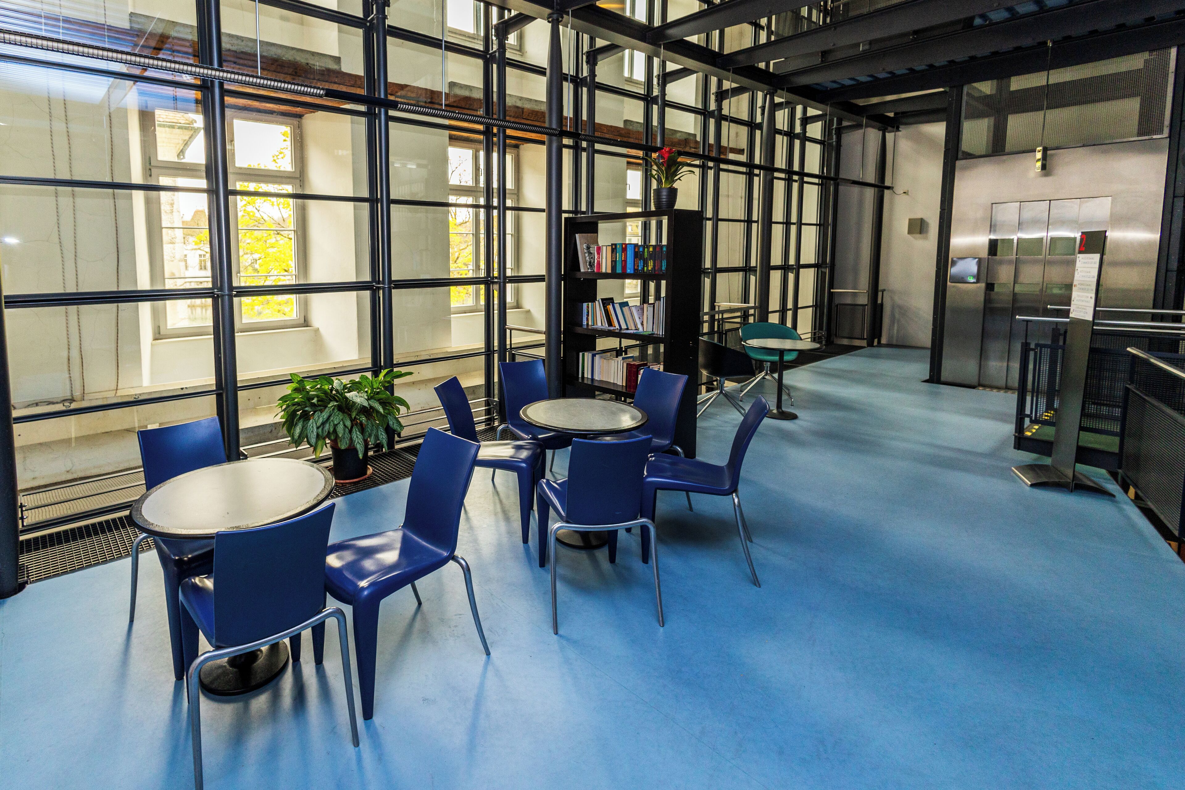 A modern indoor space with a blue floor, a round table with chairs, a bookshelf, and a potted plant.