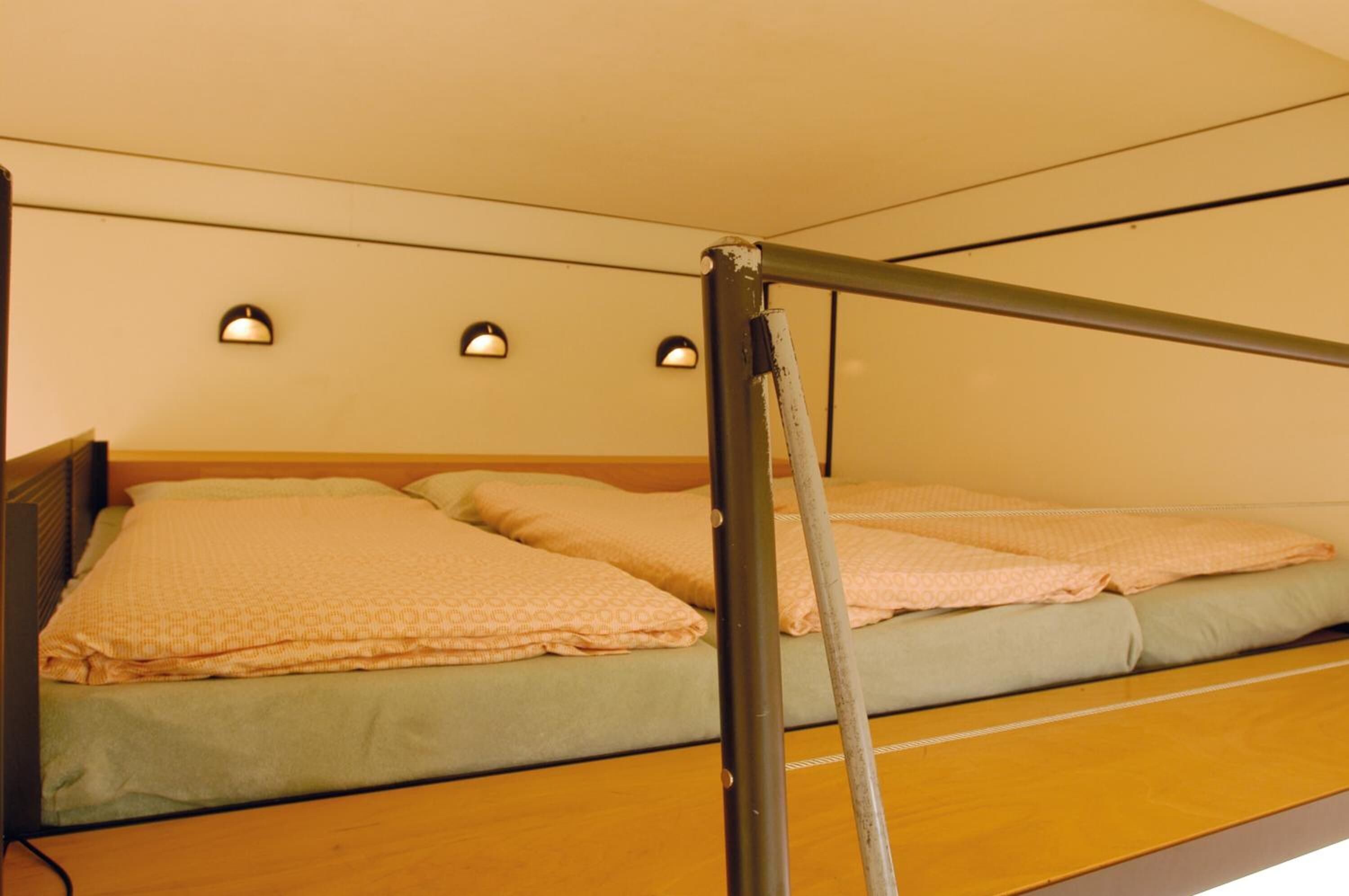 Una cama individual con cabecera de madera, estructura metálica y colchón verde. Hay tres luces fijadas a la pared sobre la cama.