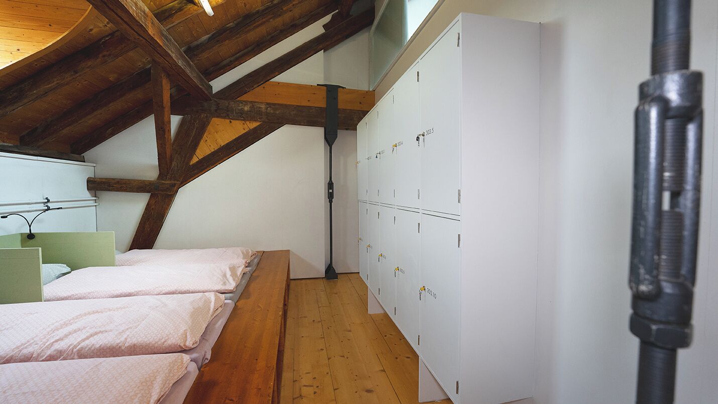 A room with wooden beams, a bed with pink bedding, and white lockers along the wall.