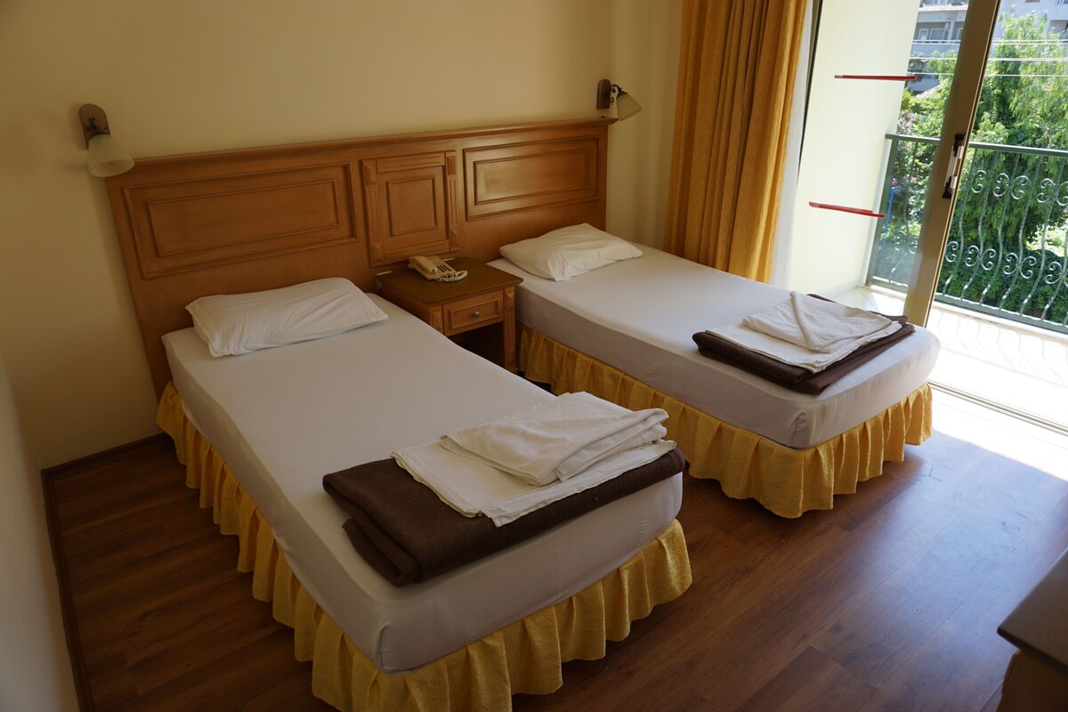 A hotel room with two beds, a nightstand, a telephone, and a window with a view of greenery.