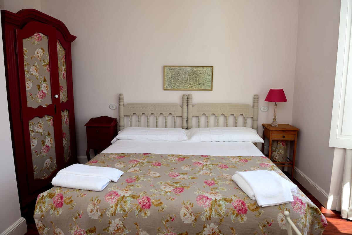 A bed with floral bedding, a wooden headboard, a red wardrobe, a nightstand with a lamp, and a framed map on the wall.