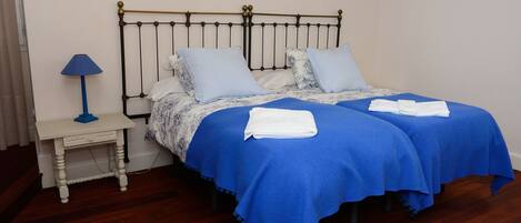 A double bed with a blue bedspread and two pillows, a small bedside table with a lamp, and a white nightstand.