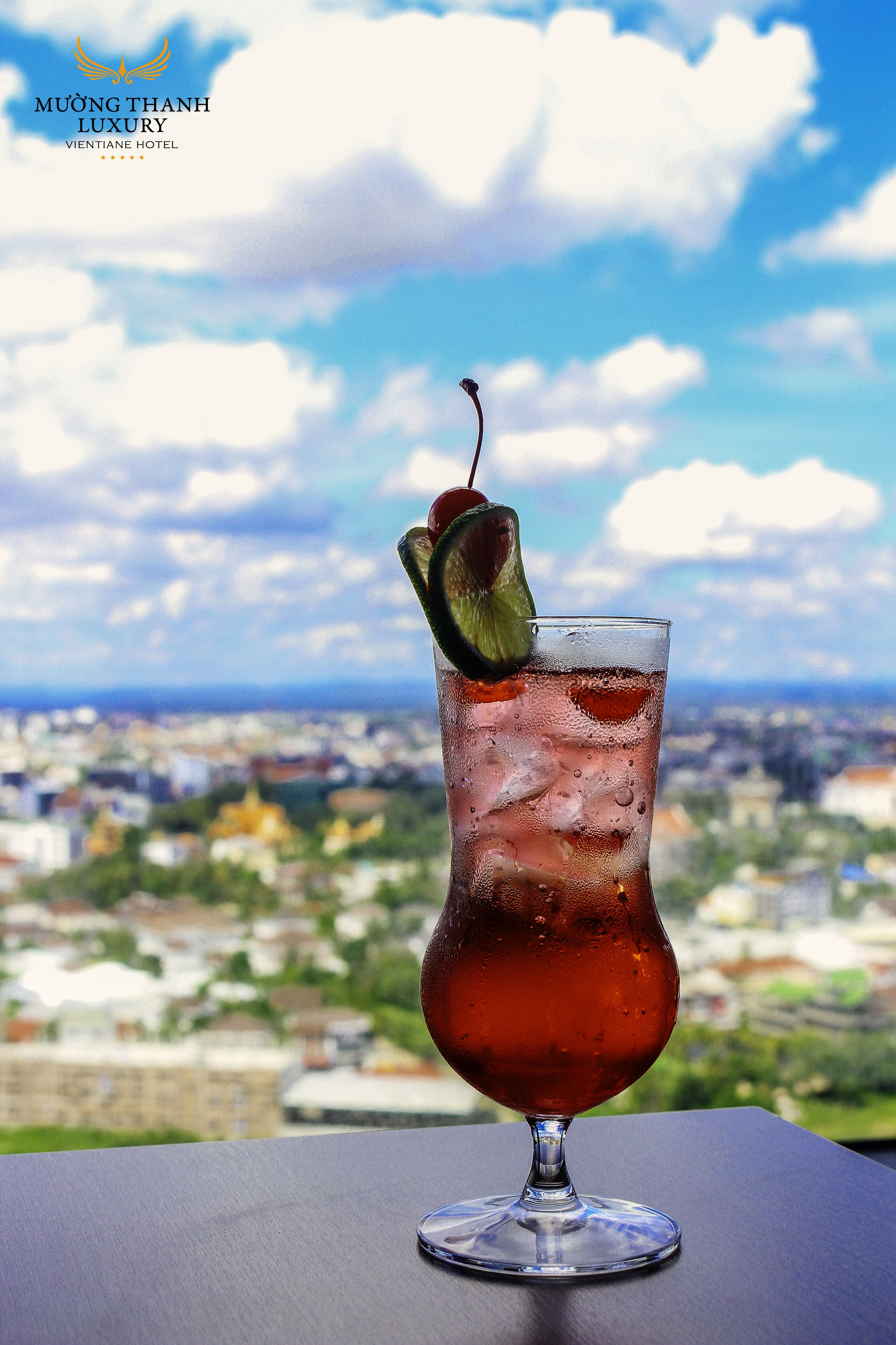 Una copa de cóctel con una cereza y lima sobre una mesa, con vista a la ciudad al fondo.