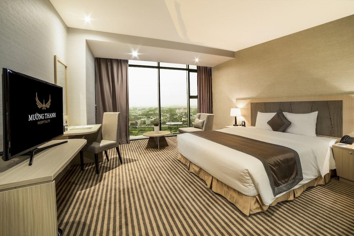 Habitación de hotel con una cama grande, un escritorio con televisor, una silla, una mesita de noche con lámpara y vistas a la ciudad.
